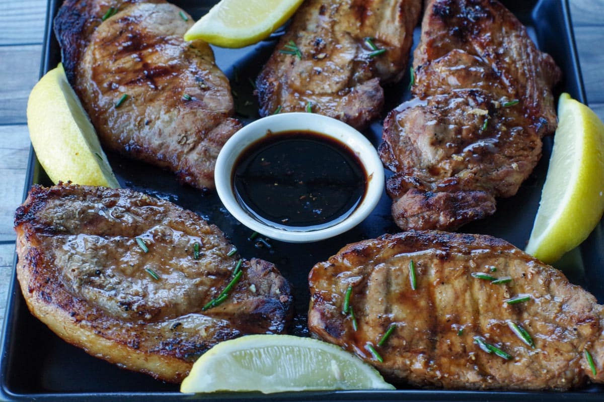 5 pieces of marinated pork on a black platter with lemon wedges and a dish of soy sauce in the middle