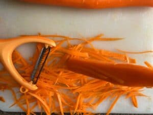 carrot being shredded with peeler on white cutting board