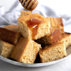 squares of cornbread piled on a plate with honey being drizzled from above