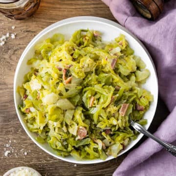 Creamed Cabbage with bacon in a white bowl on a wooden surface, with mauve linen on the side