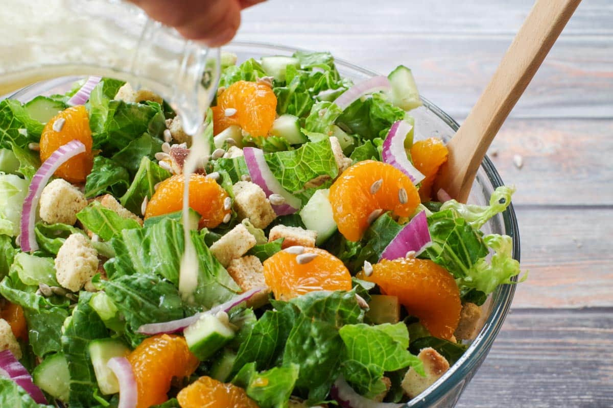 mandarin orange salad in a large glass bowl with dressing being poured on it from a glass container