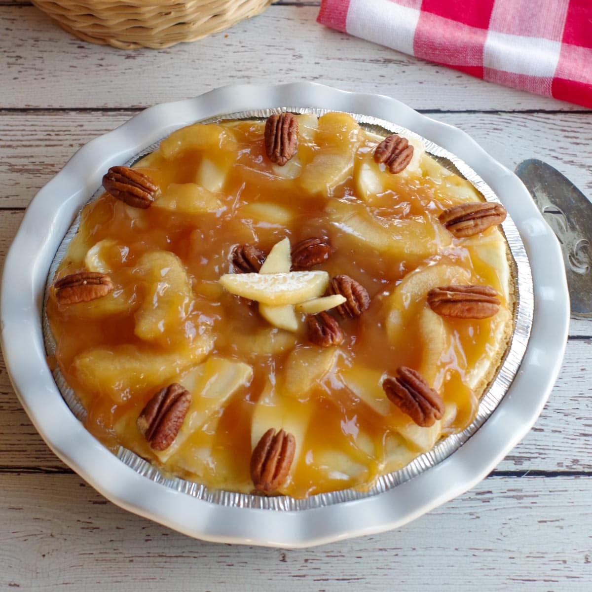 whole caramel apple cheesecake pie on a white wooden background with red checkered cloth in background