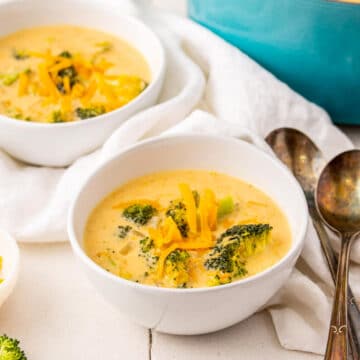 broccoli cheese soup in white bowls with white linen, spoons and blue pot in the background