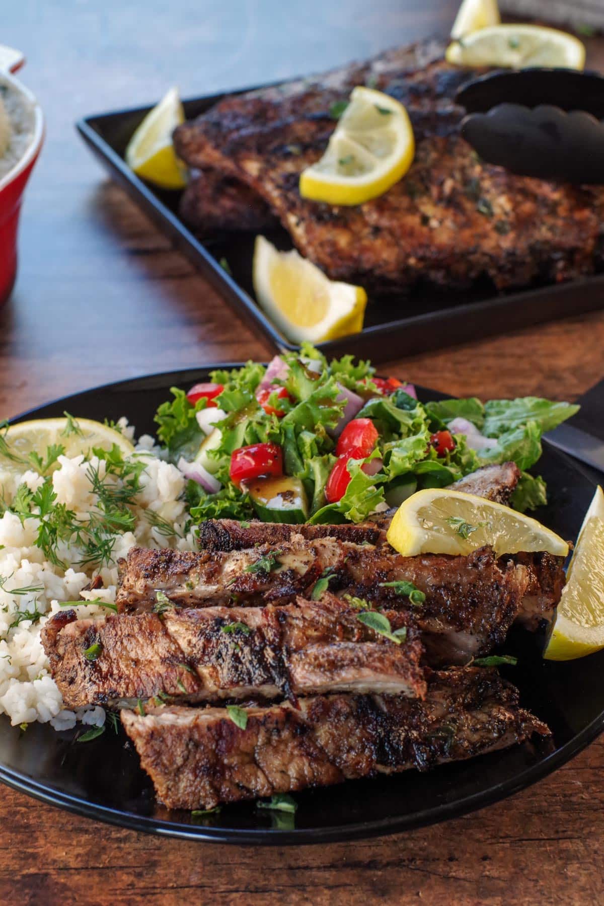 Cut Greek ribs on a plate with Greek rice and salad, with tray of ribs in the background