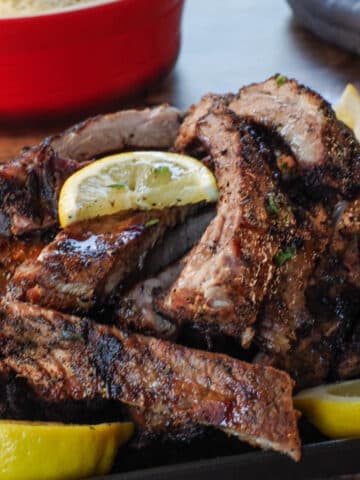Greek ribs piled on a black serving plate with lemon wedges and a red dish of rice in and grey oven mitts in the background