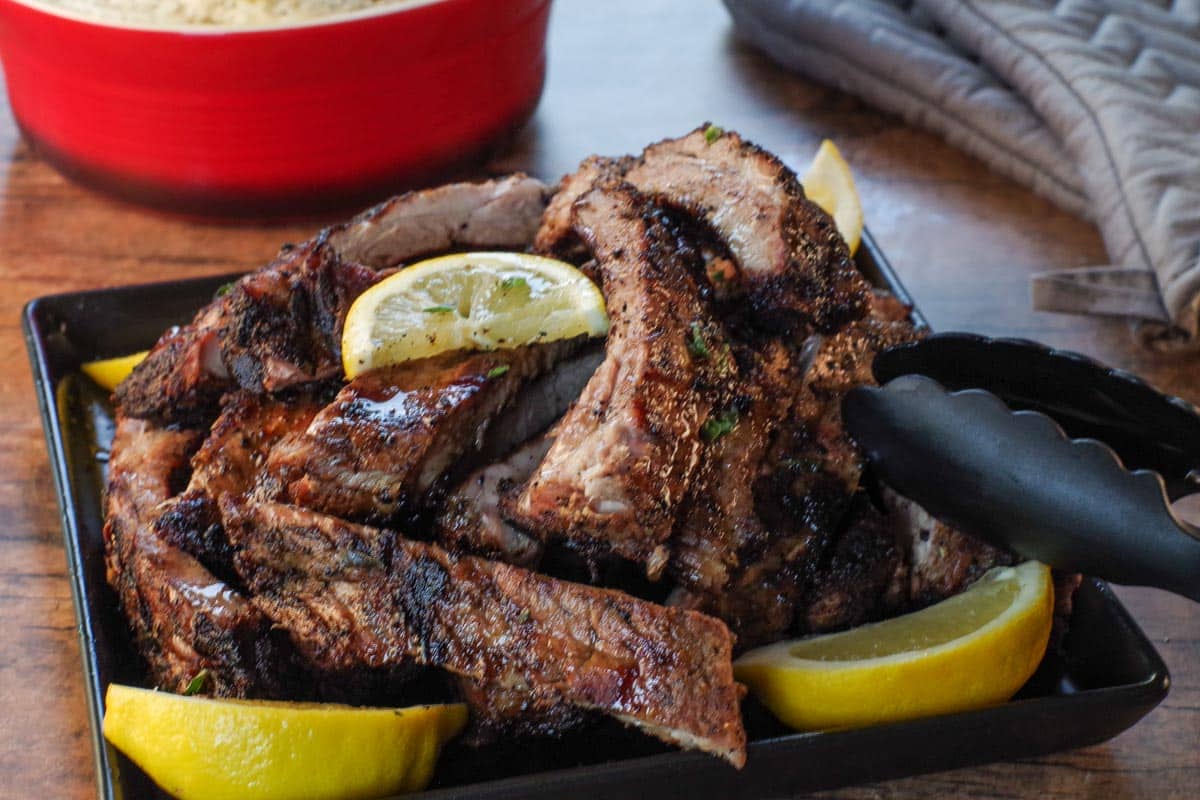 Greek ribs piled on a black serving plate with lemon wedges and a red dish of rice in and grey oven mitts in the background