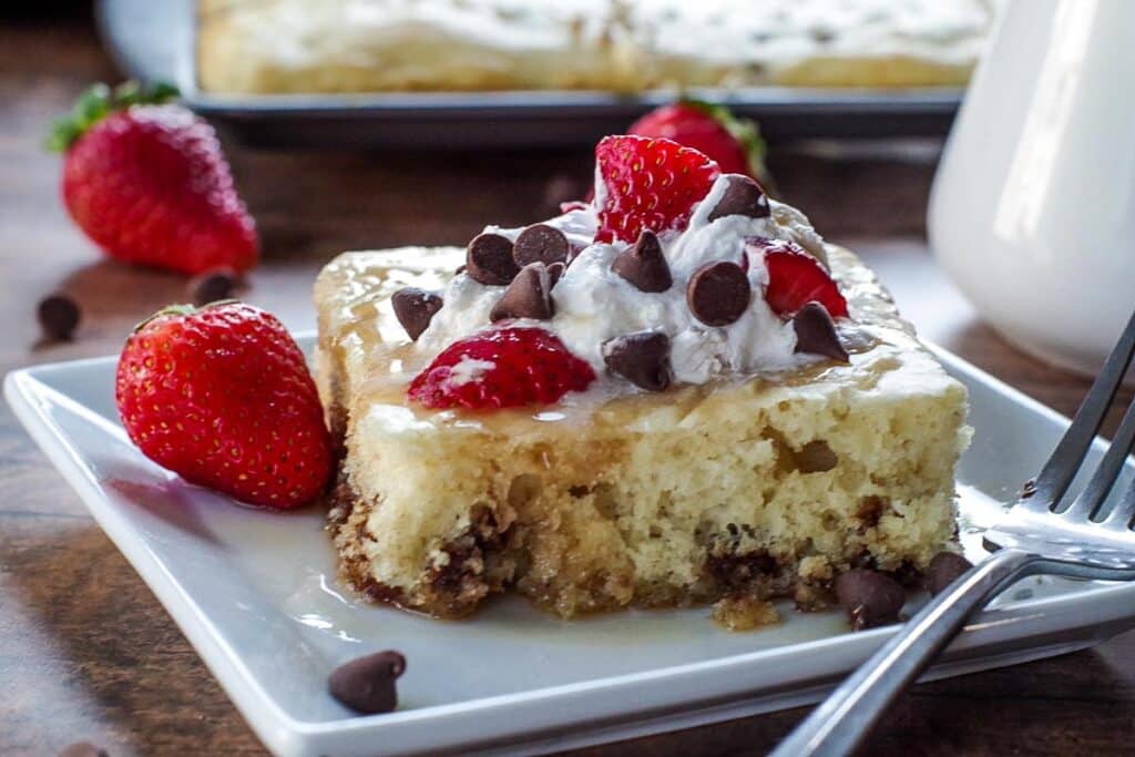 A slice of cake topped with whipped cream, fresh strawberries, and chocolate chips is on a white plate. A fork rests beside it. More strawberries and chocolate chips are scattered around, with another cake in the background.