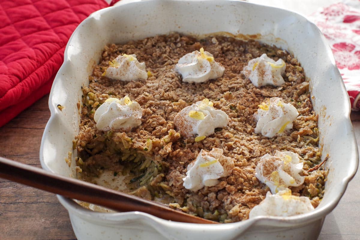 zucchini crisp in large white baking dish, dotted with whipped cream, with wooden spoon and some dessert taken out