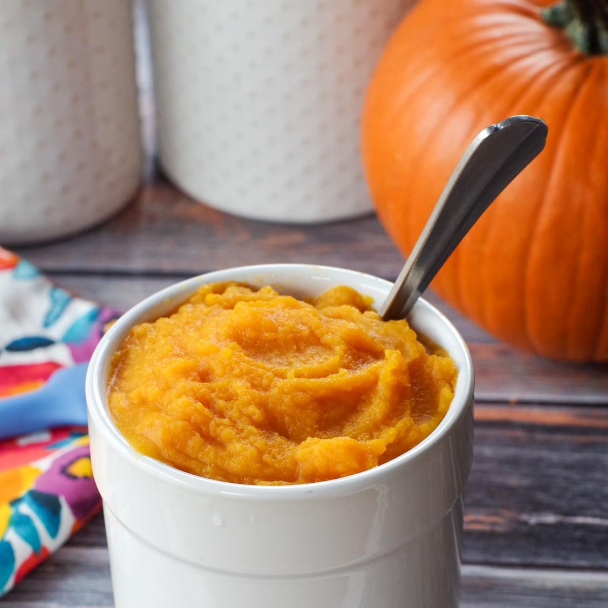 white canister with pumpkin puree in it, as well as spoon and whole pumpkin in background