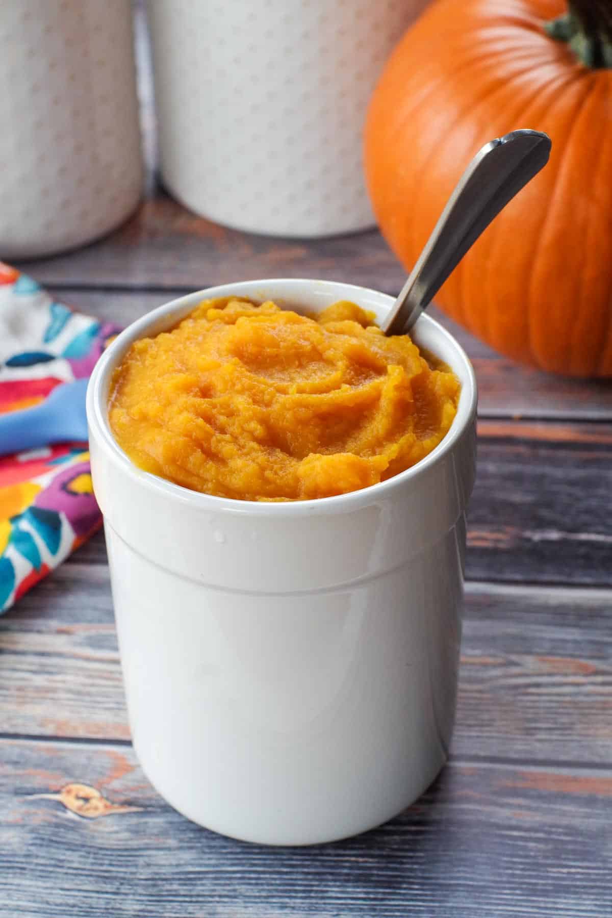 white canister with pumpkin puree in it, as well as spoon and whole pumpkin in background