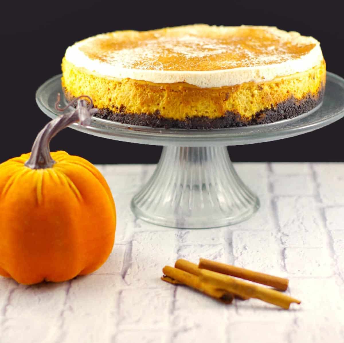 skinny pumpkin cheesecake on a glass cake stand with stuffed plush pumpkin and cinnamon sticks in front