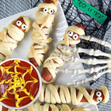 mummy hot dogs on white plate with ketchup in white bowl with spider web