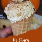 pumpkin pie ice cream in a cone, being held by a hand, in front of a container of ice cream and a pumpkin in the background
