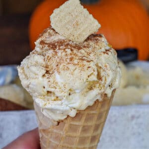 pumpkin pie ice cream in a cone, being held by a hand, in front of a container of ice cream and a pumpkin