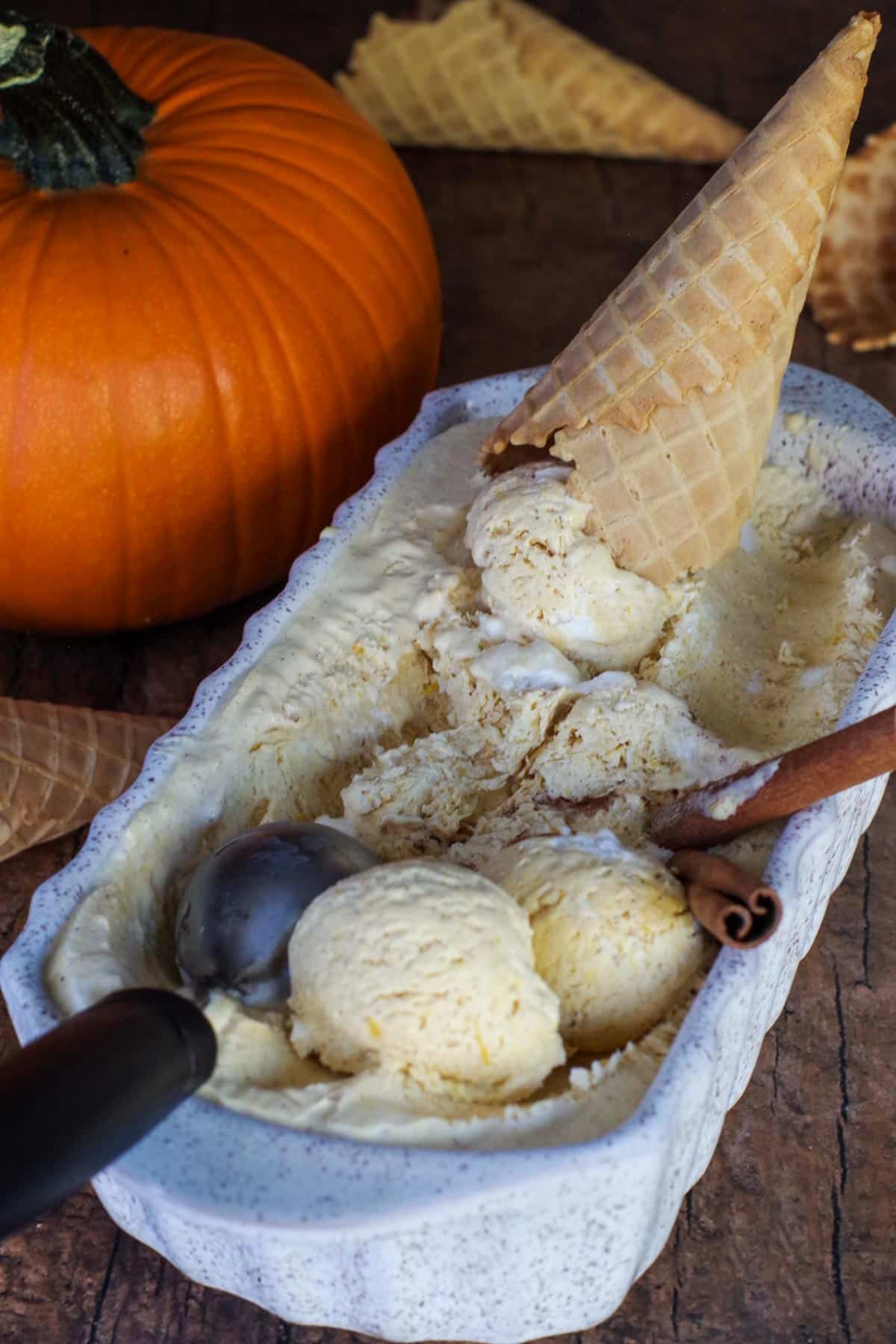 pumpkin ice cream in a beige loaf container with scooped ice cream balls, scoop, cinnamon sticks and a cone