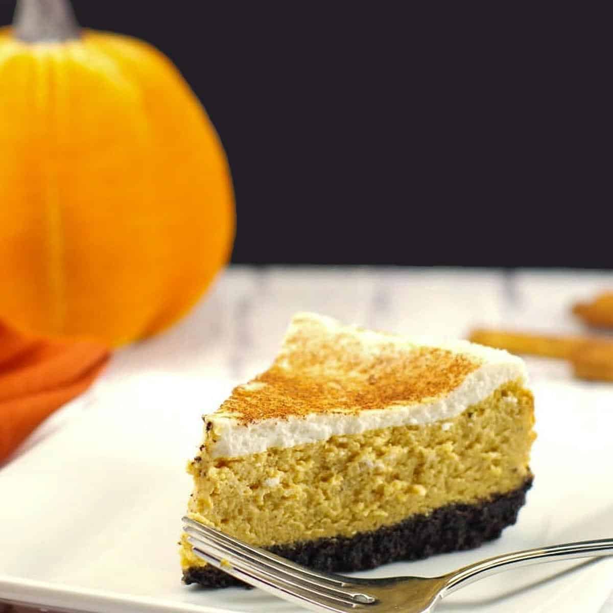 a piece of skinny pumpkin cheesecake on a white plate with fork and a stuffed pumpkin in background