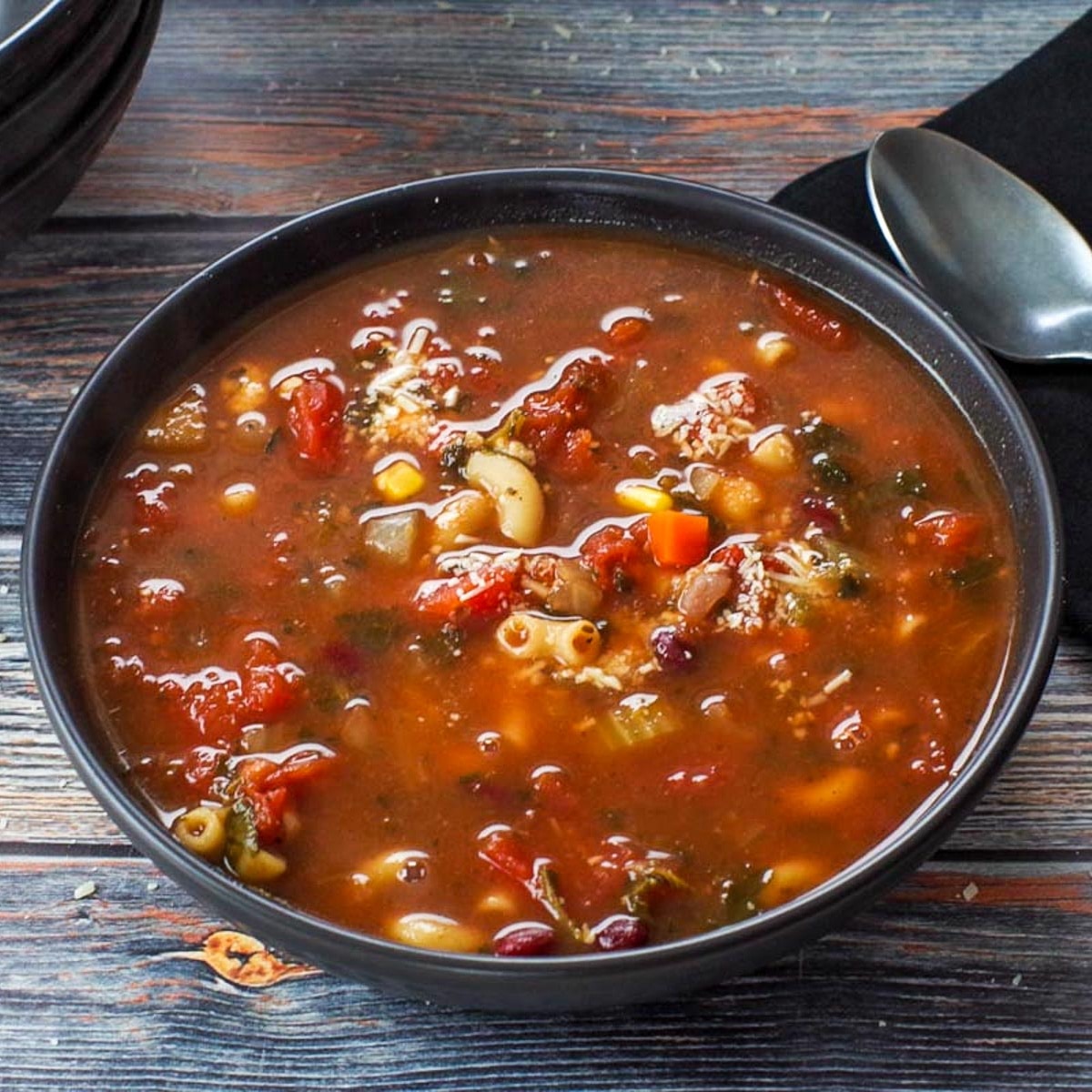 Minestrone soup in a black bowl with spoon and black napkin on the side