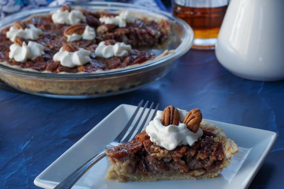 a piece of maple pecan pie on a white plate with a fork and remainder of whole pie in the background