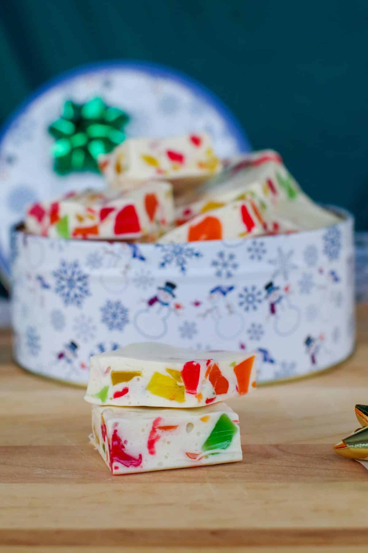 This is a photo showing a  2 square pieces Christmas Nougat stacked on top of each other with a christmas tin filled with them in the background. 