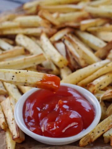 Parsnip fries on brown paper with one fry being dipped into white bowl of ketchup