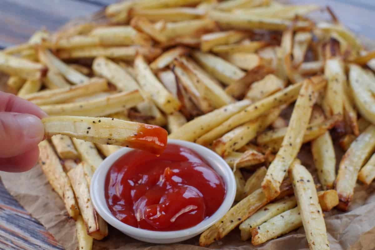 Parsnip fries on brown paper with one fry being dipped into white bowl of ketchup