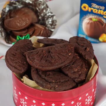 chocolate orange cookies in a red cookie tin with a chocolate orange in the back, as well as a chocolate orange box, on white material surface.