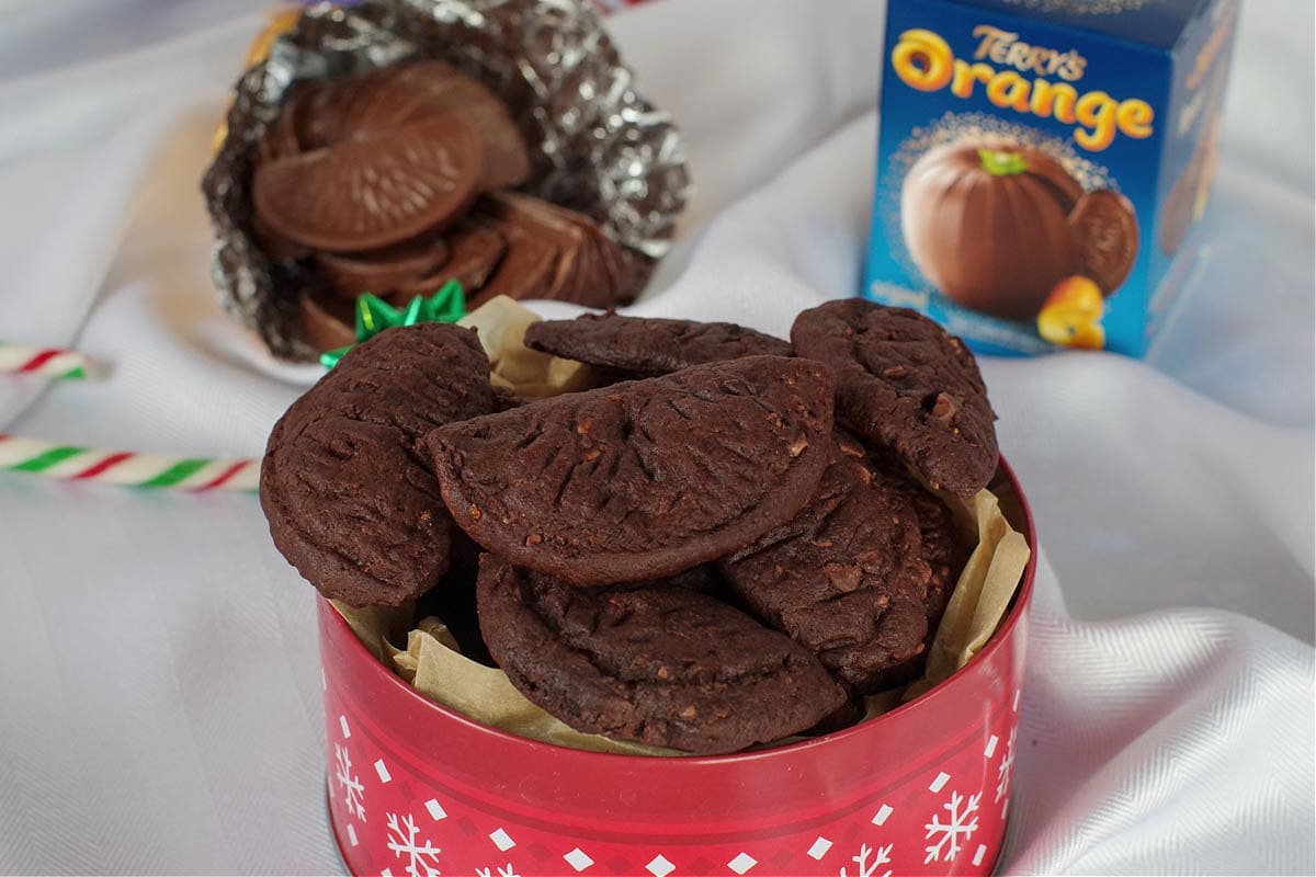 chocolate orange cookies in a red cookie tin with a chocolate orange in the back, as well as a chocolate orange box, on white material surface.