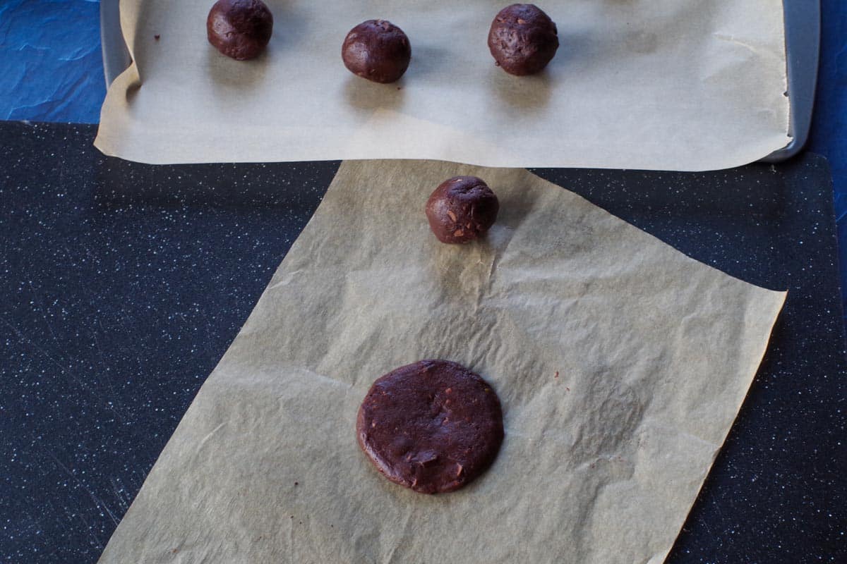 cookie dough rolled into balls on parchment paper, with one ball flattened on parchment covered black cutting board