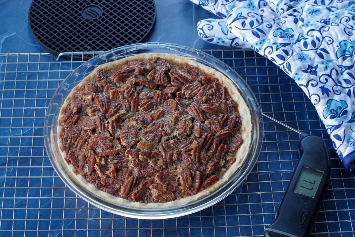 baked pecan pie on a cooling rack with black instant read thermometer beside it and black trivet and blue patterned oven mitt in the background