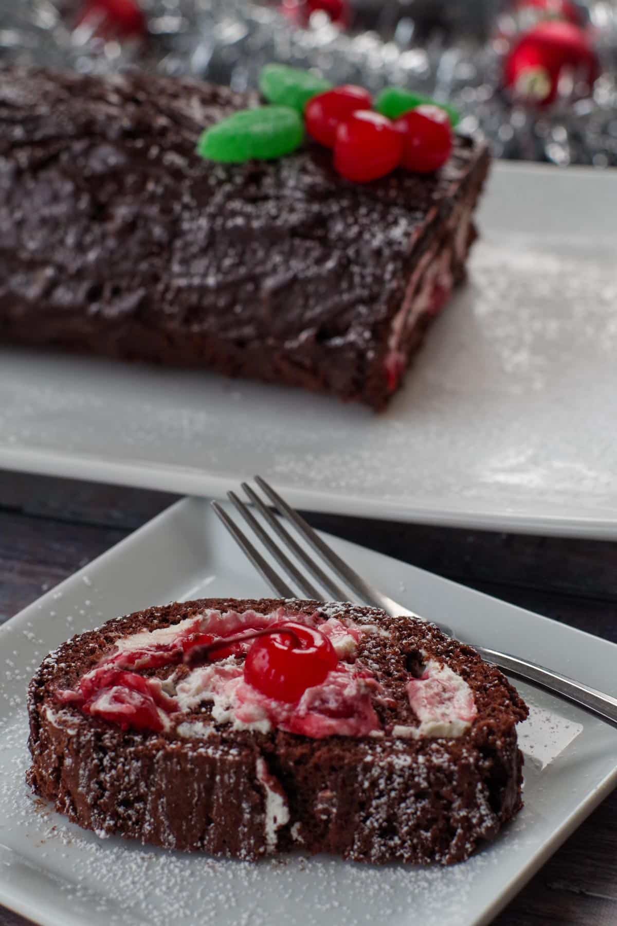 slice of black forest yule log cake on a white square plate with a fork and a whole black forest yule log cake in the background