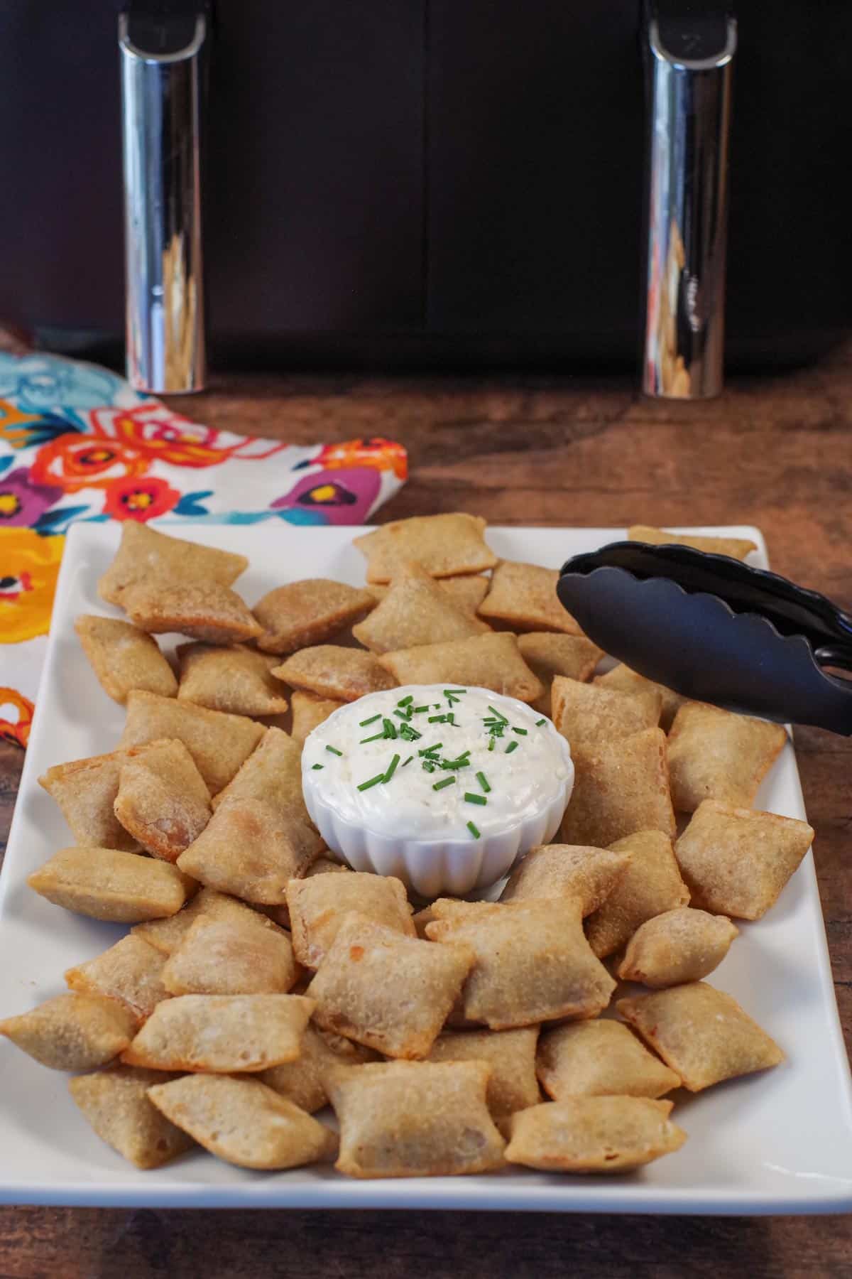 pizza bites on a white serving tray with dip in the middle and tongs on the side, and an air fryer in the background