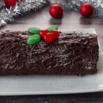 whole black forest yule log cake on a white platter with tinsel and red tree decorations in background