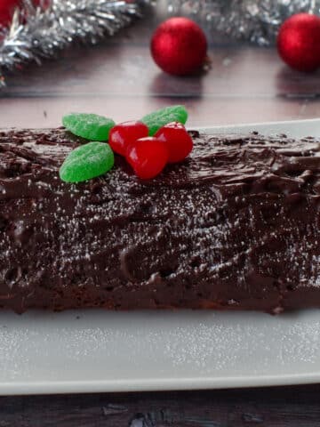 whole black forest yule log cake on a white platter with tinsel and red tree decorations in background