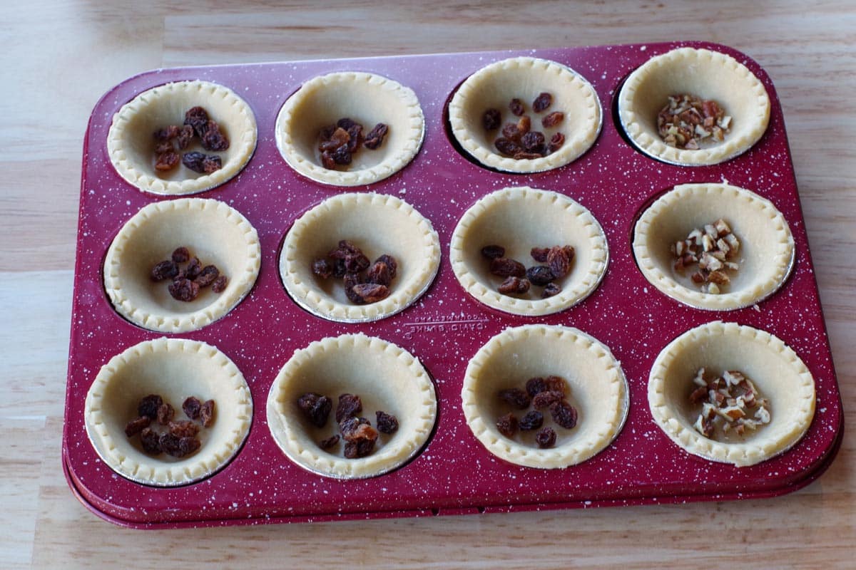 tarts shells in red speckled muffin tin with raisins and pecans in bottom of tarts