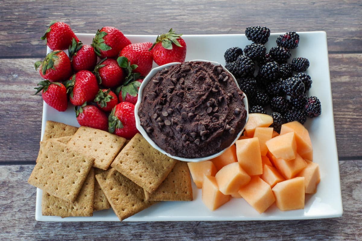 chocolate dessert hummus in a white petal bowl, on a white platter with fruit and graham crackers