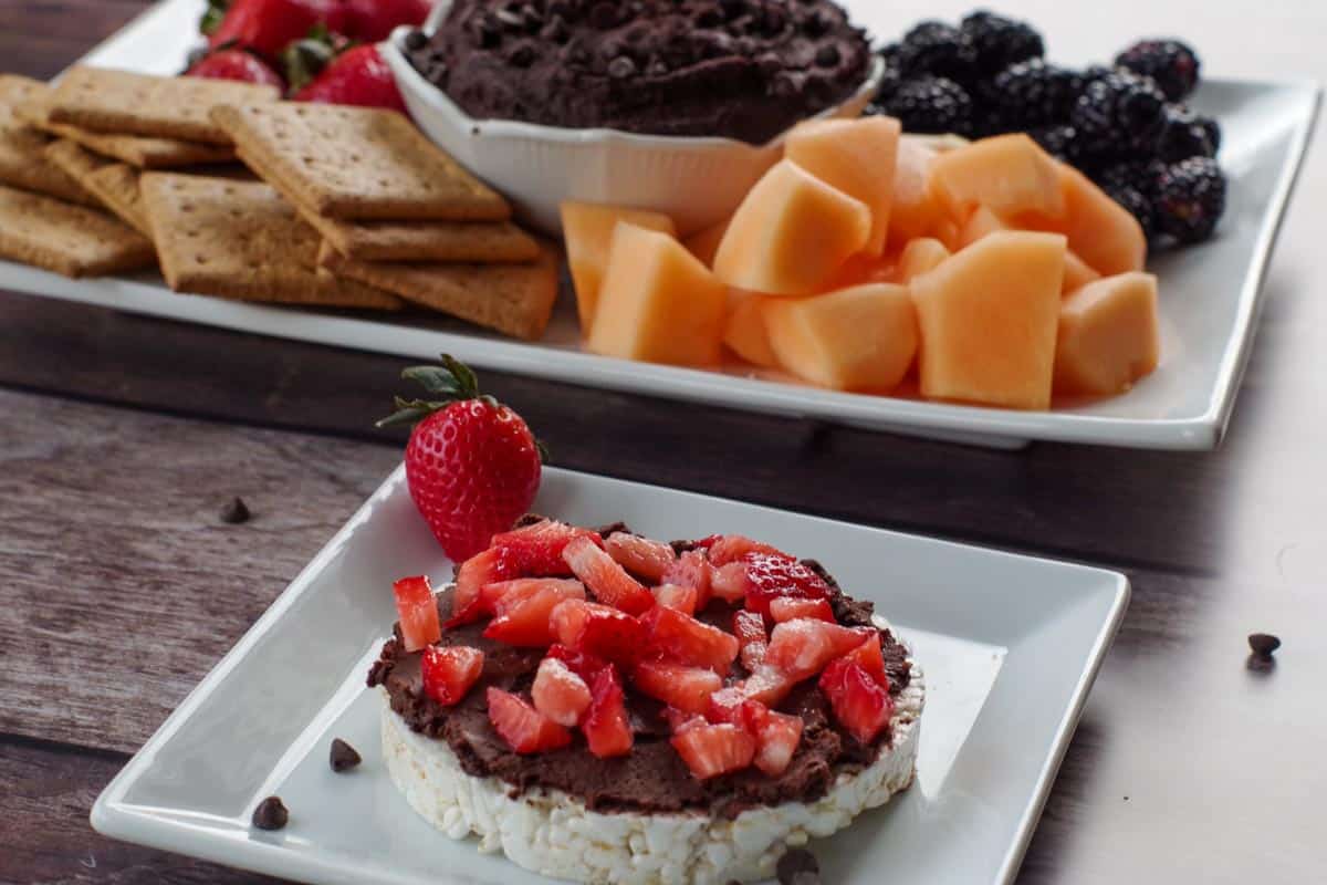 chocolate hummus on a rice cake with strawberries and platter of hummus and fruit in background