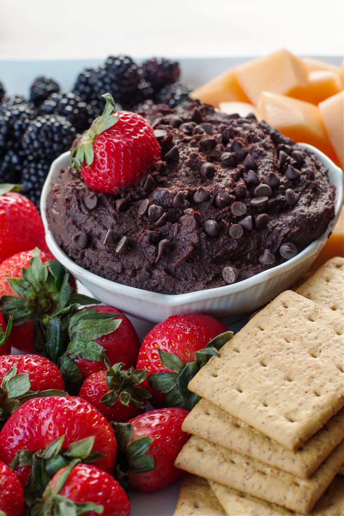 Dark Chocolate Hummus in a whit petal bowl, with a strawberry in the hummus, on a platter of fruit and graham crackers