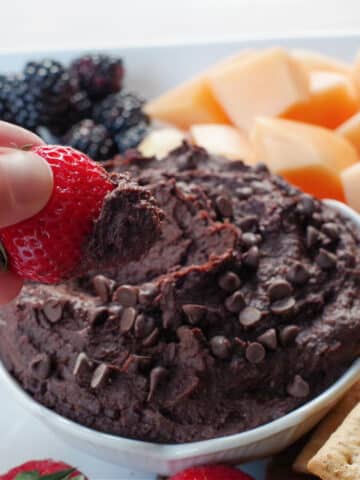 dark chocolate hummus in a white petal bowl (on a white platter with fruit and crackers), with a strawberry being dipped into it