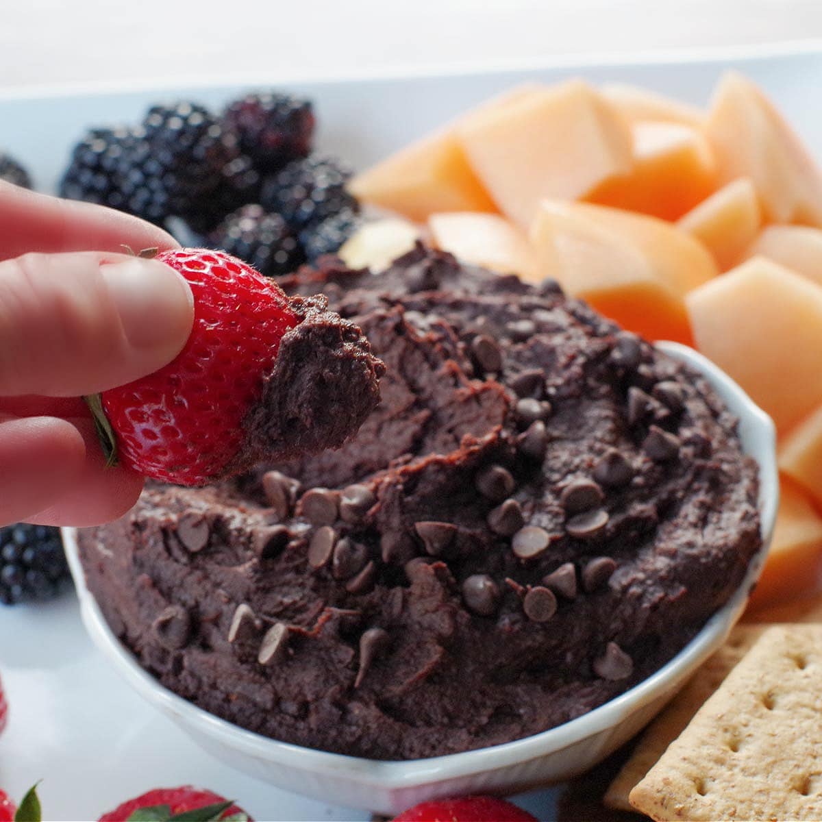 dark chocolate hummus in a white petal bowl (on a white platter with fruit and crackers), with a strawberry being dipped into it