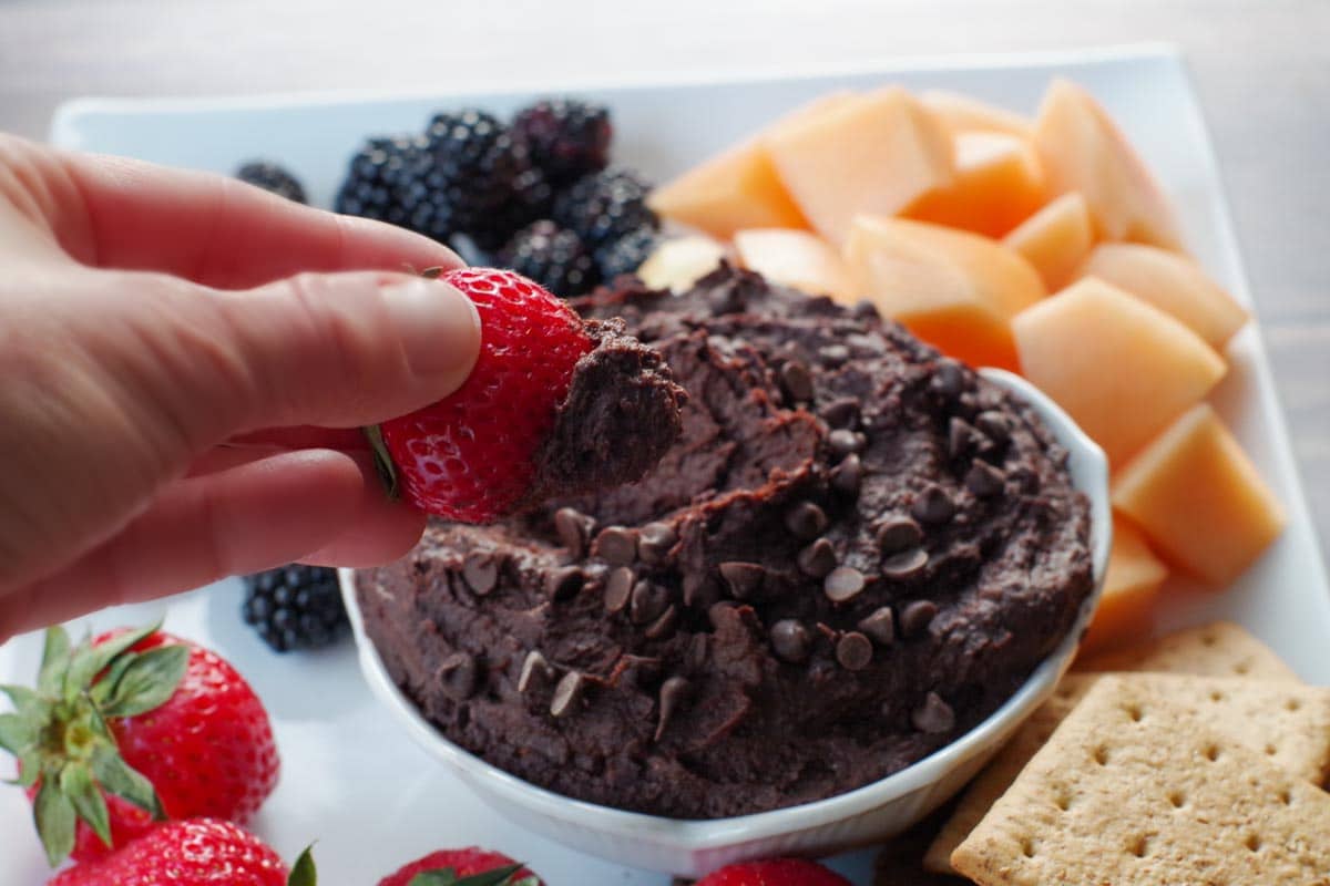 dark chocolate hummus in a white petal bowl (on a white platter with fruit and crackers), with a strawberry being dipped into it