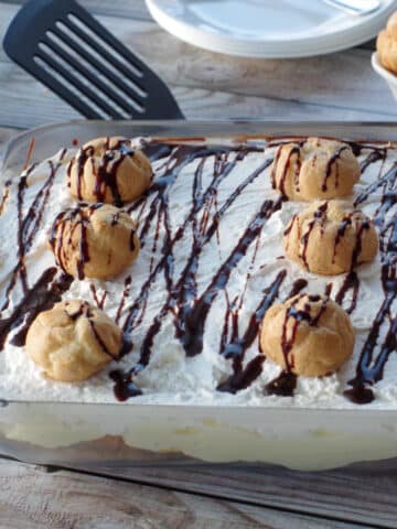 cream puff dessert in a glass dish with plates and a bowl of cream puffs in background