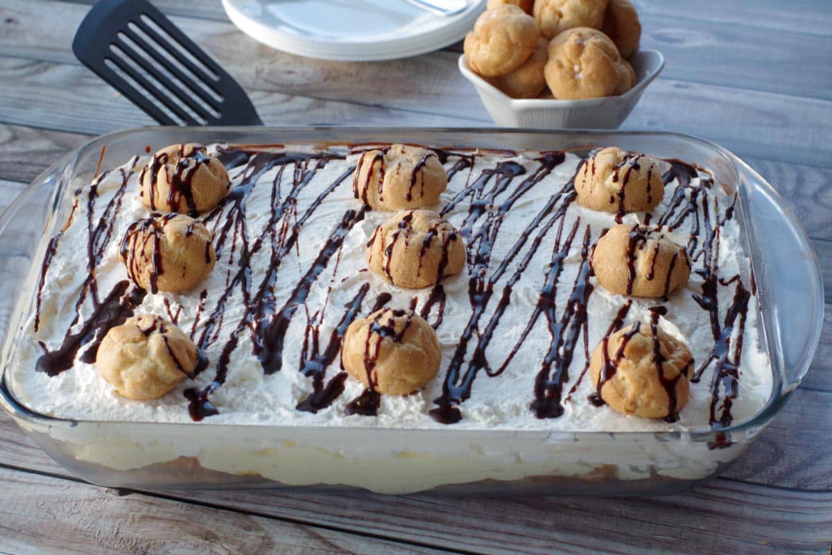 cream puff dessert in a glass dish with plates and a bowl of cream puffs in background