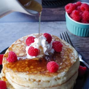 3 pancakes with raspberry, syrup and whipped cream stacked on a black plate with syrup being poured over them