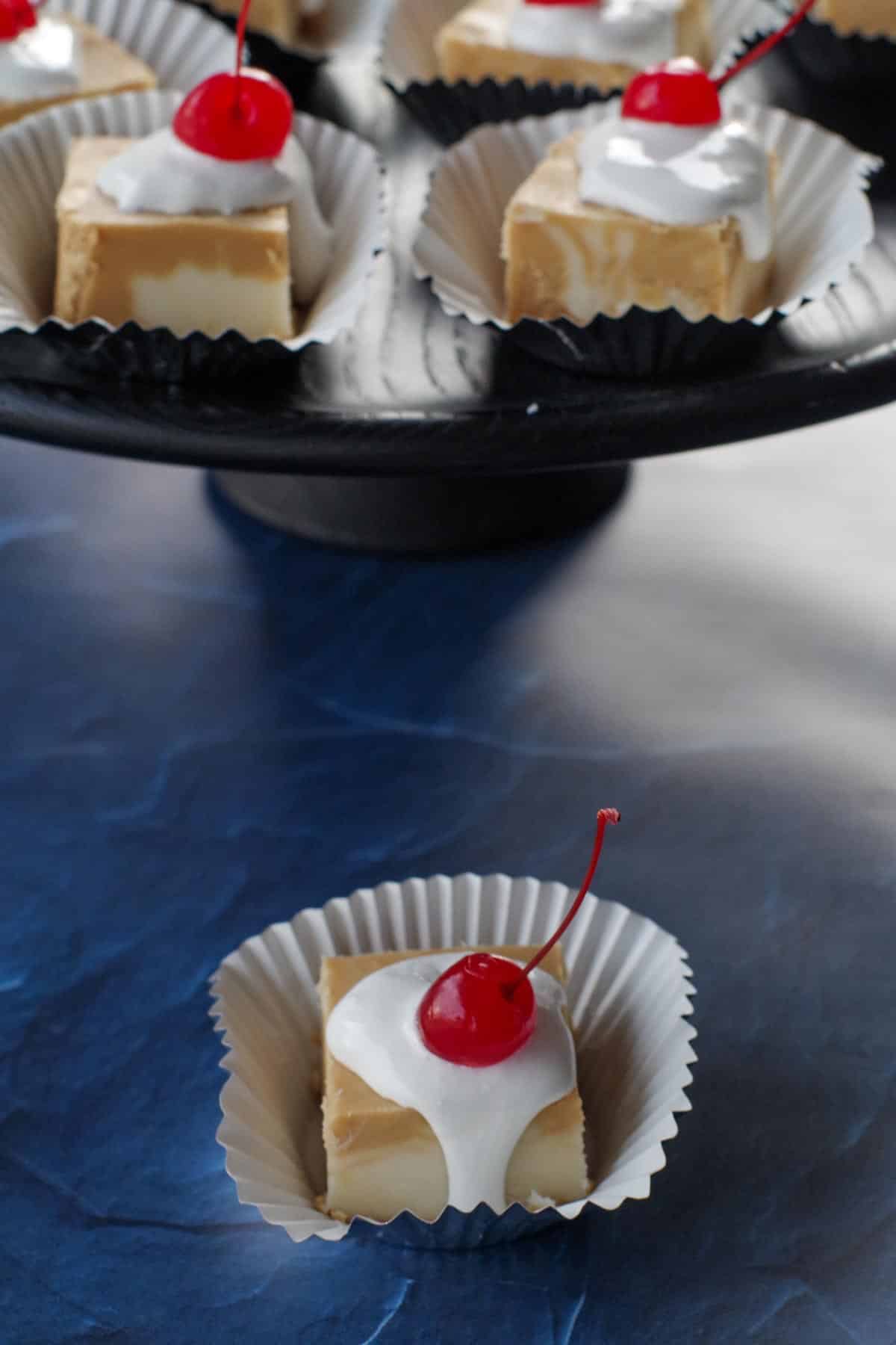 a piece of root beer float fudge on a cupcake liner, in front of a black tray with more root beer float fudge pieces