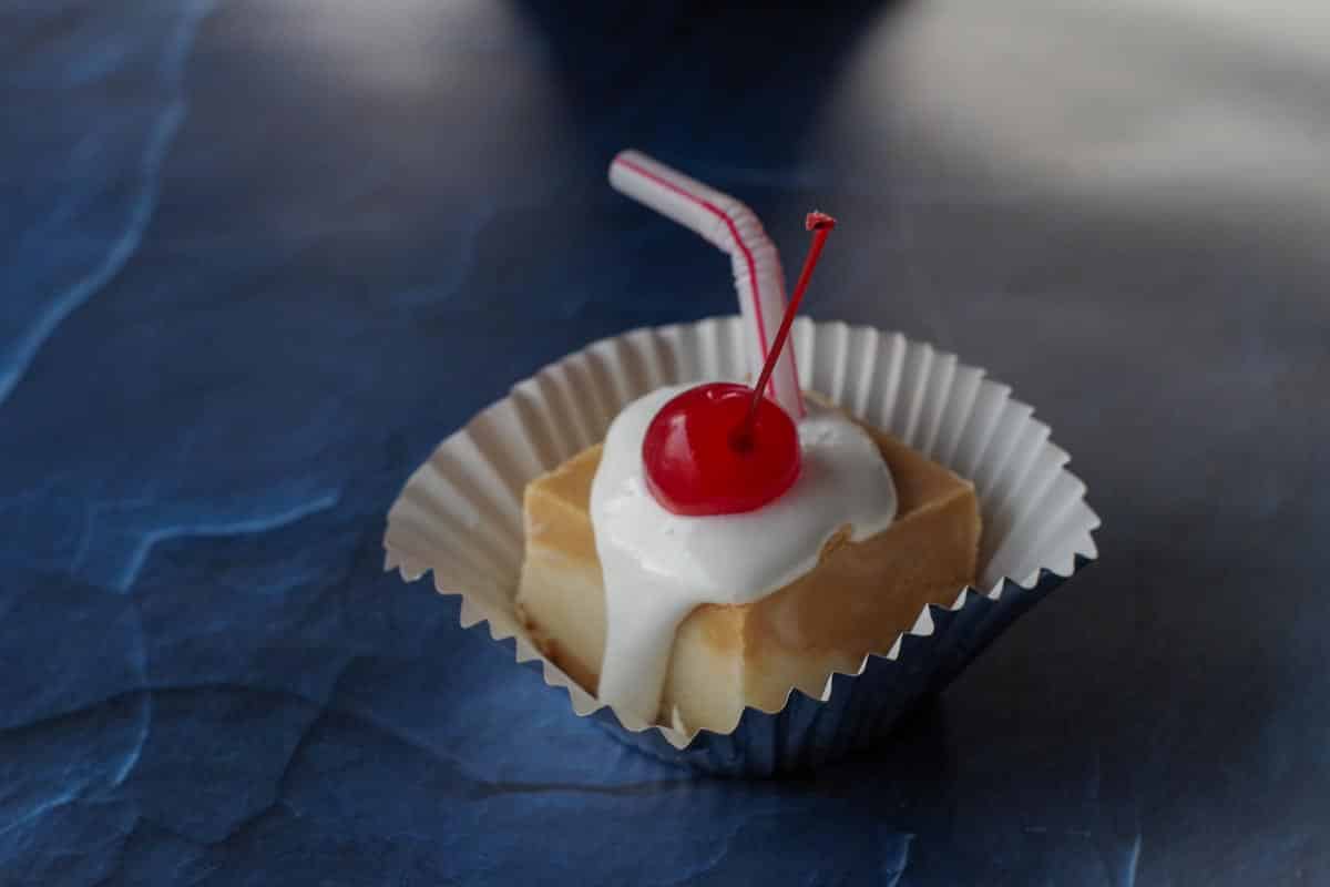 a piece a root beer float fudge on a cupcake liner, with marshmallow fluff and cherry, with a straw in it on a blue surface