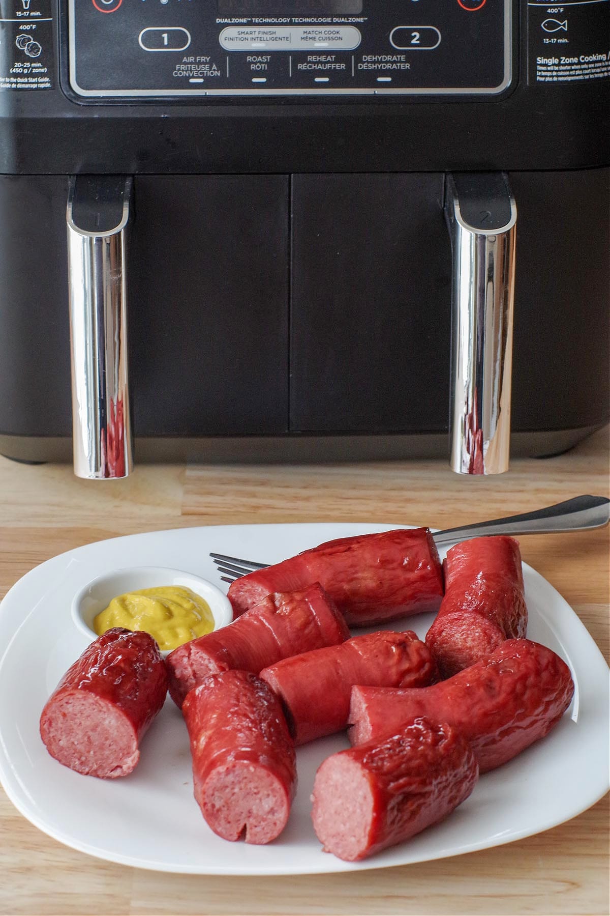 kielbasa on a white plate with a small white dish of mustard and tongs, with an air fryer in the background