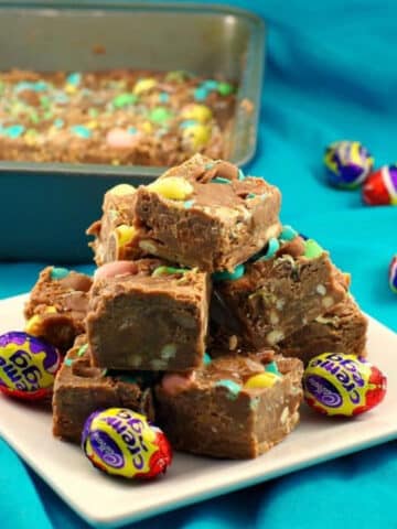 Squares of Easter Creme egg fudge piled on a white plate with a pan of fudge in the background