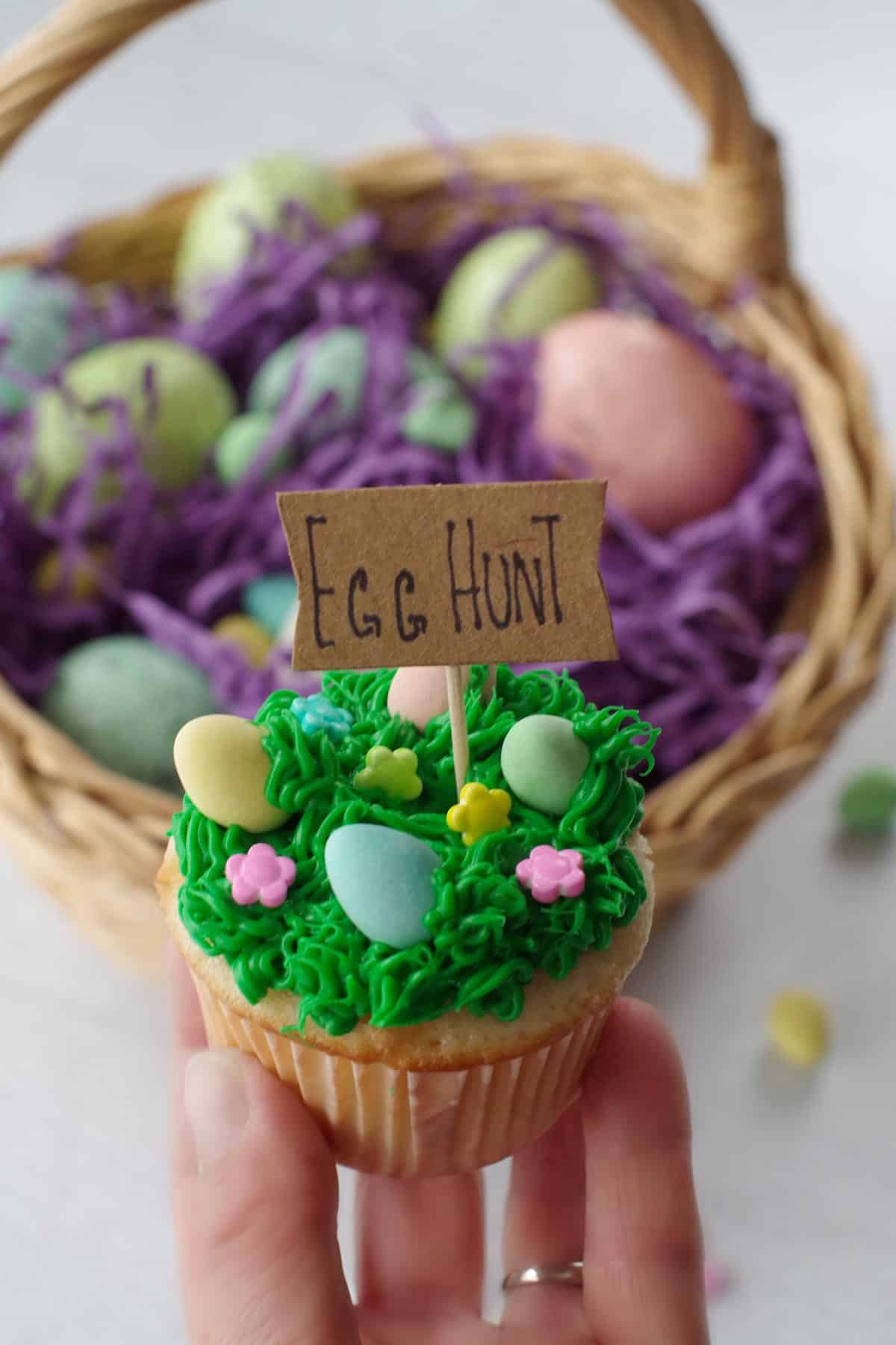 Easter Mini Egg cupcake being held up in front of an Easter egg basket.