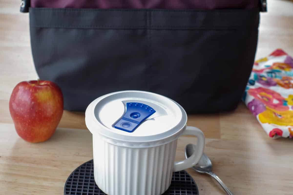 Individual pot pie in a white meal mug with a lid, in front of lunch bag, with apple in the background