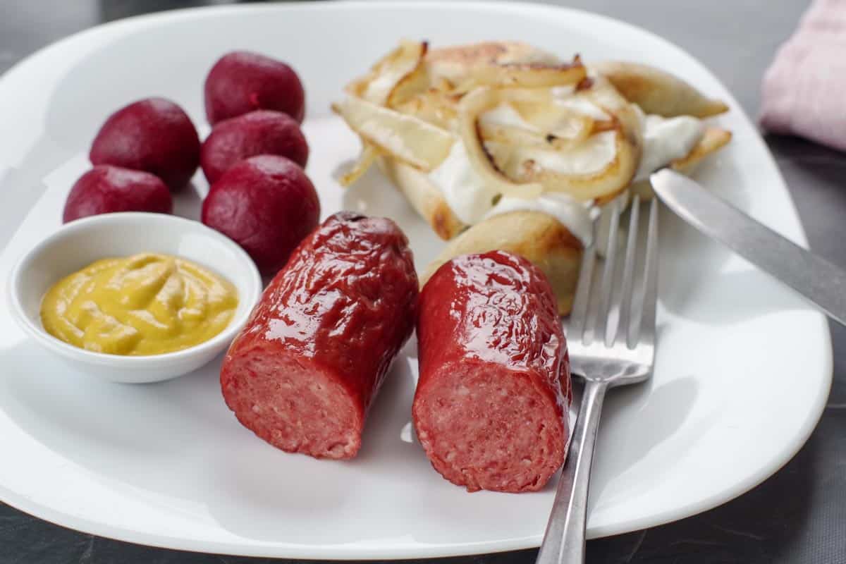 2 pieces of kielbasa with a small dish of mustard and beets and perogies on a white plate, with a fork